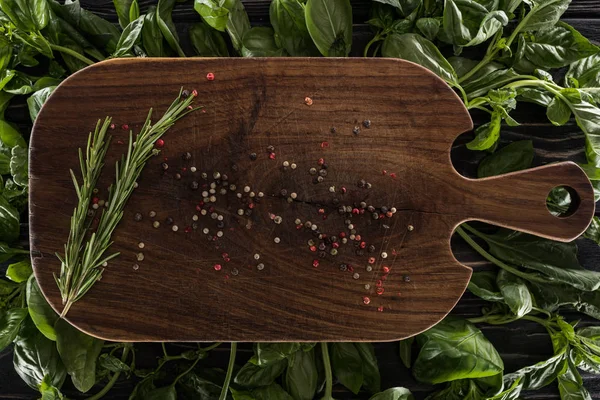 Vista superior de tabla de cortar de madera con especias y romero sobre hojas de albahaca - foto de stock