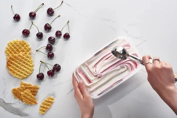 Vista recortada de la mujer con delicioso helado y cuchara cerca de gofres y cerezas sobre fondo gris mármol - foto de stock