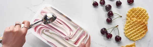 Vista recortada de la mujer con delicioso helado y cuchara cerca de gofres y cerezas sobre fondo gris mármol, tiro panorámico - foto de stock