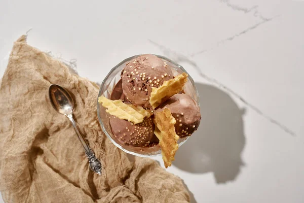 Top view of delicious chocolate ice cream in bowl with waffles near brown cloth with spoon on marble grey background — Stock Photo