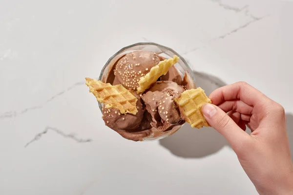 Vista cortada de mulher segurando waffle perto de delicioso sorvete de chocolate na tigela no fundo cinza mármore — Fotografia de Stock