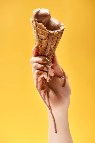 Vista parcial de la mujer sosteniendo delicioso helado de chocolate derretido en cono de gofre crujiente aislado en amarillo - foto de stock