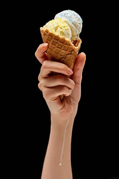 Vista recortada de la mujer que sostiene el derretimiento delicioso helado de vainilla con aspersiones en cono de gofre crujiente aislado en negro - foto de stock