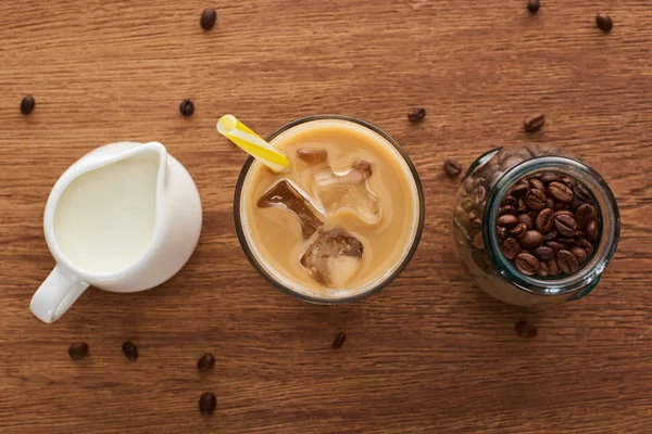 Vue du dessus du café glacé avec de la paille près du lait dans une cruche et des grains de café dans un pot sur une table en bois — Photo de stock
