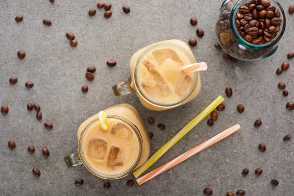 Top view of ice coffee in glass jars near straws and coffee grains on grey background — Stock Photo