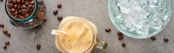 Top view of ice coffee in glass jar with straw near coffee grains and ice cubes on grey background, panoramic shot — Stock Photo