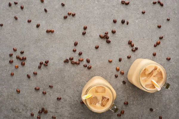 Top view of ice coffee in glass jars with straws and scattered coffee grains on grey background — Stock Photo