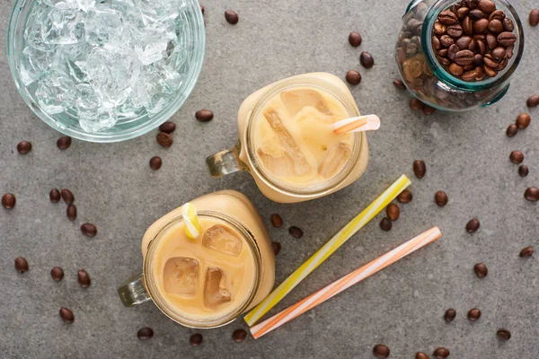 Vista superior del café helado en frascos de vidrio cerca de pajitas, granos de café y cubitos de hielo sobre fondo gris - foto de stock