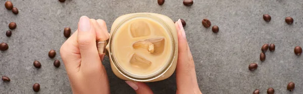 Cropped view of woman holding glass jar with ice coffee and coffee grains on grey background, panoramic shot — Stock Photo