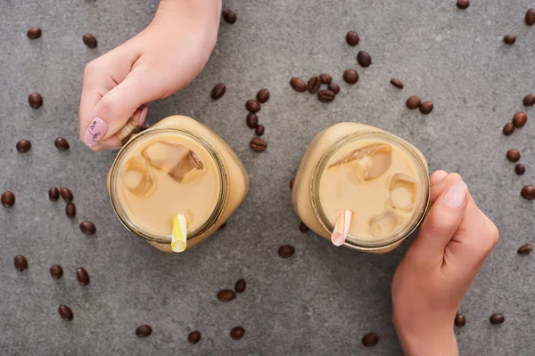 Vista recortada de las mujeres sosteniendo frascos de vidrio con granos de café y café sobre fondo gris - foto de stock