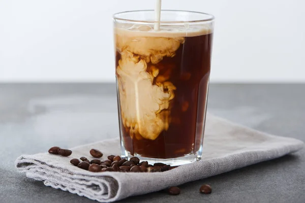 Leche vertida en el café helado en vaso sobre servilleta con granos de café aislados en blanco - foto de stock