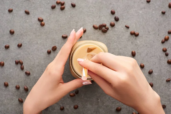Vue partielle de la femme tenant un bocal en verre avec paille et café glacé près des grains de café sur fond gris — Photo de stock