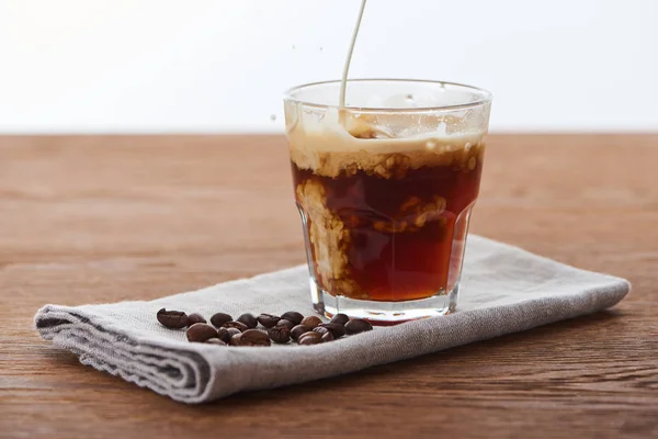 Milk pouring into ice coffee in glass on napkin with coffee grains on wooden table isolated on white — Stock Photo