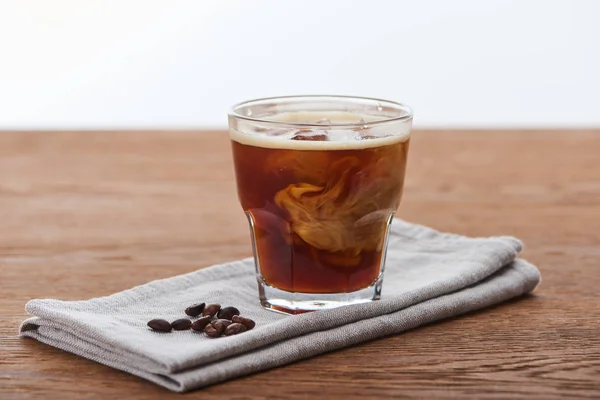 Ice coffee mixing with milk in glass on napkin with fesh coffee grains on wooden table isolated on white — Stock Photo