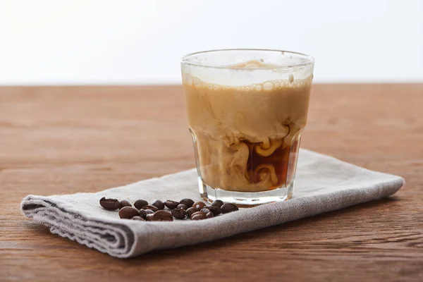 Ice coffee mixing with milk in glass on napkin with coffee grains on wooden table isolated on white — Stock Photo