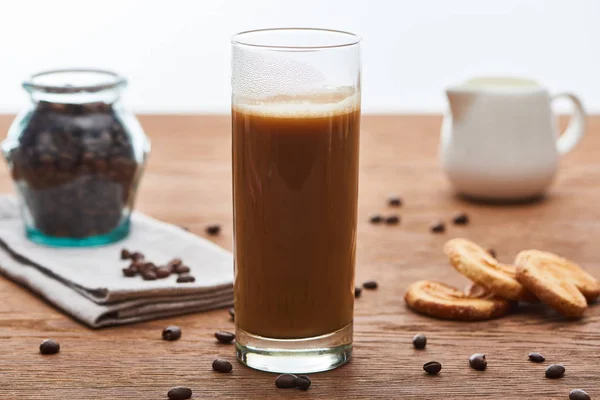 Selective focus of ice coffee with milk in glass near milk jug, cookies and coffee grains on wooden table isolated on white — Stock Photo