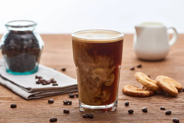 Messa a fuoco selettiva della miscelazione del caffè ghiacciato con latte in vetro vicino alla brocca del latte, biscotti e chicchi di caffè su tavolo di legno isolato su bianco — Foto stock