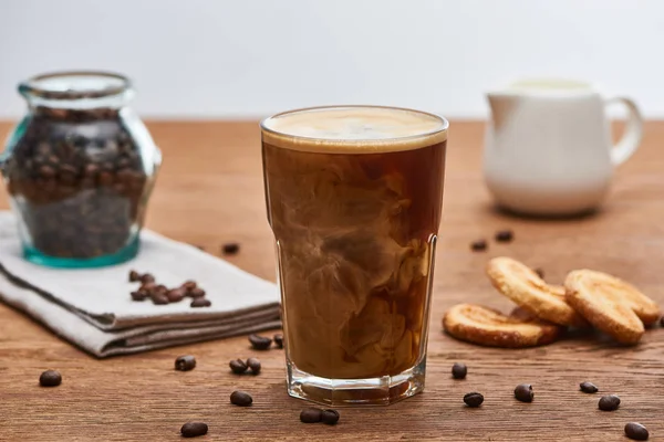 Messa a fuoco selettiva della miscelazione del caffè ghiacciato con latte in brocca di vetro vicino al latte, biscotti e chicchi di caffè su tavolo di legno isolato su grigio — Foto stock