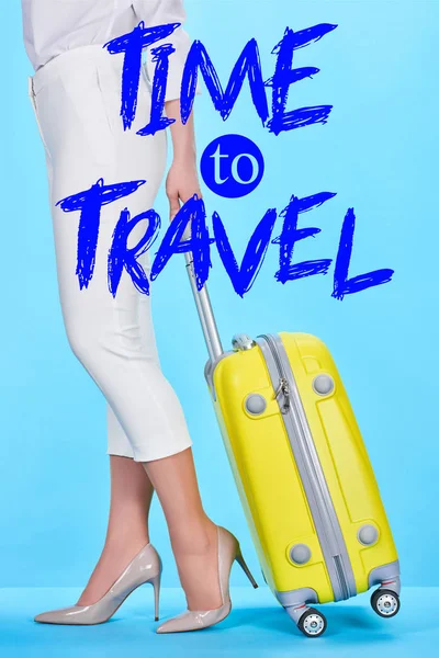 Cropped view of woman holding handle of yellow travel bag on blue background with time to travel illustration — Stock Photo