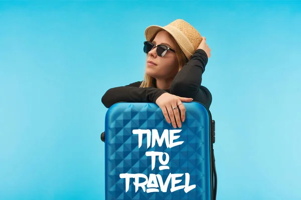 Блондинка в сонцезахисних окулярах і солом'яному капелюсі біля синьої валізи з часом для подорожей ілюстрація ізольована на синьому — стокове фото