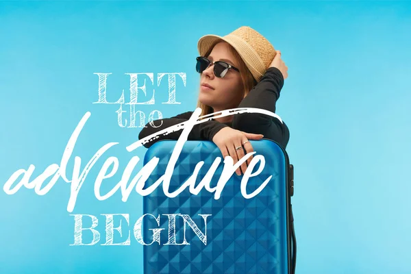 Menina loira em óculos de sol e chapéu de palha perto de mala azul isolada em azul com deixe a aventura começar ilustração — Fotografia de Stock
