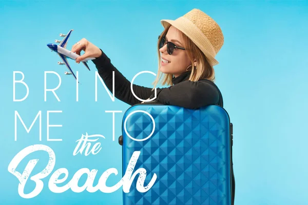 Блондинка щаслива дівчина в сонцезахисних окулярах і солом'яній обшивці капелюха з іграшковим літаком біля синьої сумки для подорожей ізольована на синьому з приводом мене на пляж ілюстрація — стокове фото