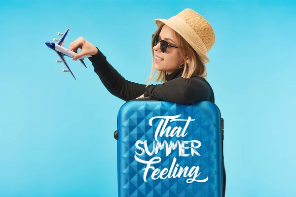 Rubia chica feliz en gafas de sol y sombrero de paja chapado con avión de juguete cerca de la bolsa de viaje azul con esa sensación de verano letras aisladas en azul - foto de stock