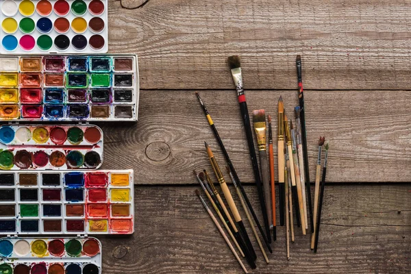 Vue de dessus des palettes de peinture colorées sur la surface brune en bois avec pinceaux — Photo de stock