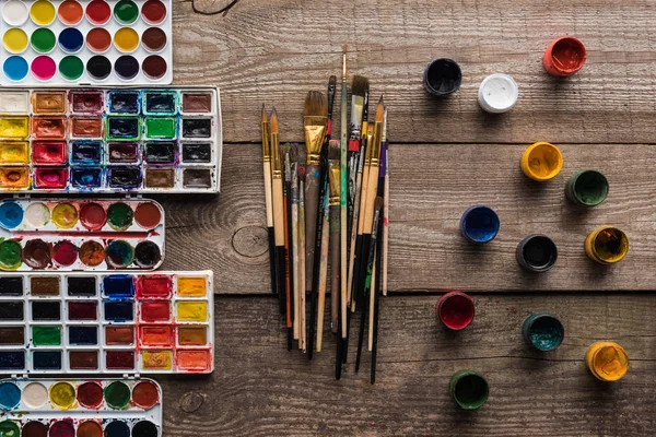 Top view of colorful paint palettes on wooden brown surface with paintbrushes and gouache — Stock Photo