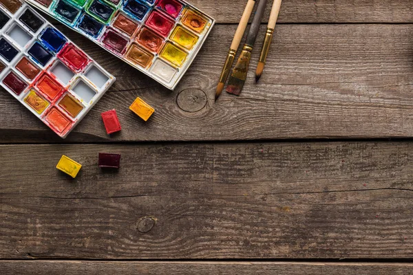 Vista dall'alto di tavolozze di colori ad acquerello e pennelli su superficie di legno con spazio copia — Foto stock