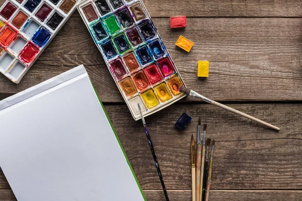 Top view of colorful paint palettes, paintbrushes and blank sketch pad on wooden surface — Stock Photo