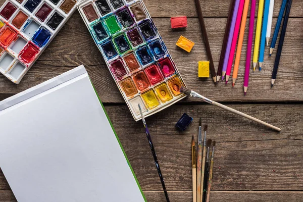 Top view of colorful paint palettes, paintbrushes, color pencils and blank sketch pad on wooden surface — Stock Photo