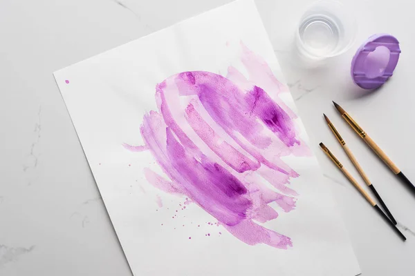 Top view of purple watercolor brushstrokes on white paper near paintbrushes on marble white surface — Stock Photo