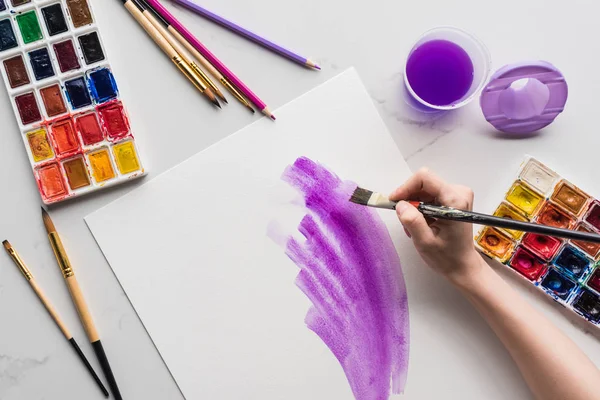 Vista ritagliata di artista disegno pennellate acquerello viola su carta bianca su superficie di marmo bianco — Foto stock