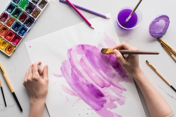 Частичный вид художника, рисующего фиолетовые акварельные мазки на белой бумаге на мраморной белой поверхности — стоковое фото
