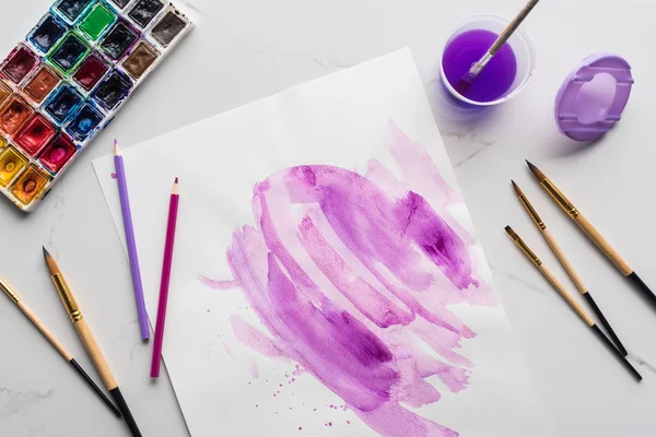 Вид зверху фіолетові акварельні мазки на білому папері біля пензлів, фарб та кольорових олівців на мармуровій білій поверхні — стокове фото