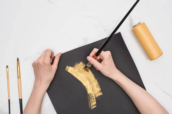 Cropped view of artist drawing with golden paint in black paper on marble surface — Stock Photo