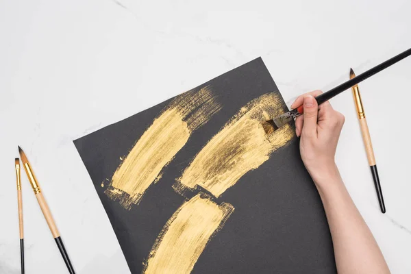 Partial view of artist drawing brushstrokes with golden paint in black paper on marble surface — Stock Photo