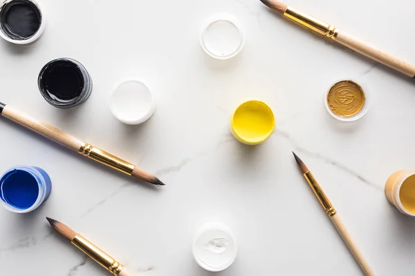 Top view of colorful gouache paints and scattered paintbrushes on marble white surface — Stock Photo