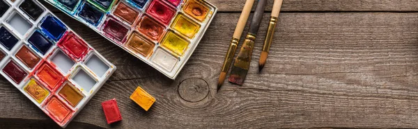 Vista dall'alto di tavolozze di colori ad acquerello su superficie marrone legno con pennelli, colpo panoramico — Foto stock