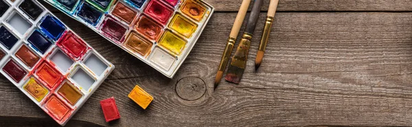 Vue de dessus des palettes de peinture à l'aquarelle sur surface marron bois avec pinceaux, vue panoramique — Photo de stock