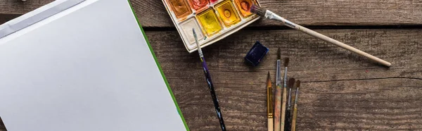 Top view of colorful paint palette, paintbrushes and blank sketch pad on wooden surface, panoramic shot — Stock Photo