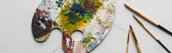 Top view of colorful palette with oil paints near paintbrushes on marble white surface, panoramic shot — Stock Photo