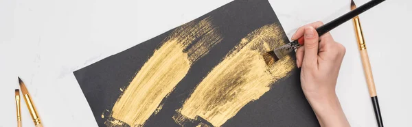Partial view of artist drawing brushstrokes with golden paint in black paper on marble surface, panoramic shot — Stock Photo