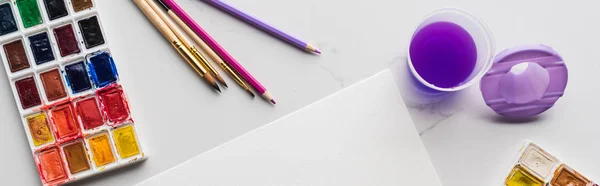 Вид зверху на порожній білий папір біля кольорових олівців та акварельних фарб на мармуровій білій поверхні, панорамний знімок — стокове фото