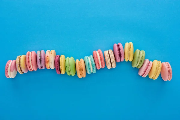 Vista superior de macarrones franceses deliciosos multicolores en línea curva sobre fondo azul brillante - foto de stock