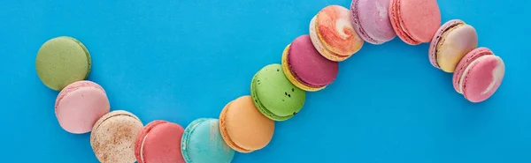 Vista superior de multicoloridos deliciosos macaroons franceses em linha curva no fundo azul brilhante com espaço de cópia, tiro panorâmico — Fotografia de Stock