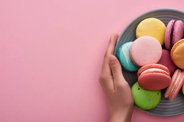 Cropped view of woman holding plate with multicolored delicious French macaroons on pink background with copy space — Stock Photo