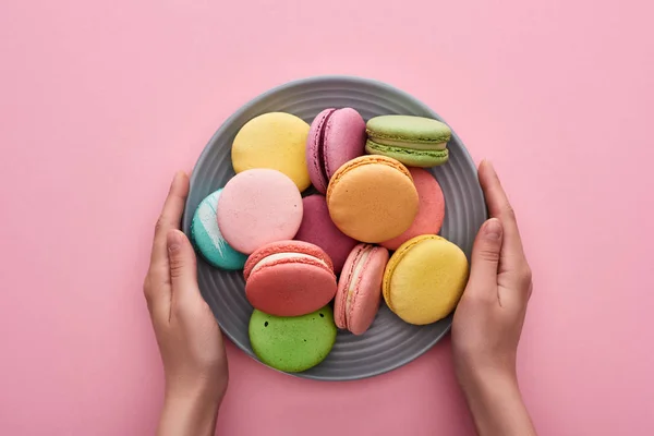 Vista cortada da mulher segurando placa com macaroons franceses deliciosos multicoloridos no fundo rosa — Fotografia de Stock