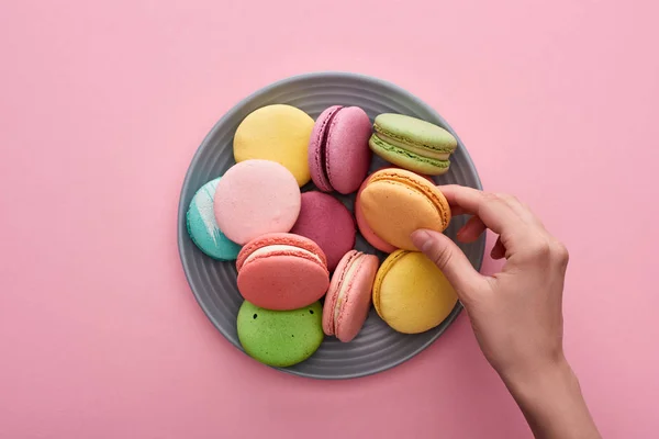 Cropped view of female hand near plate with multicolored delicious French macaroons on pink background — Stock Photo